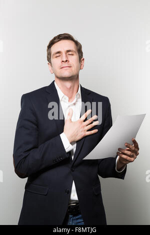 Blond-haired businessman puts his hand on his chest and feels a touching moment while in front of a gradient background Stock Photo