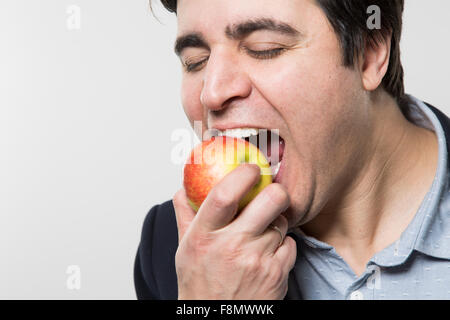 Dark-haired european businessman with a cheerful look takes a bite out of an small apple with his eyes closed while in front of  Stock Photo