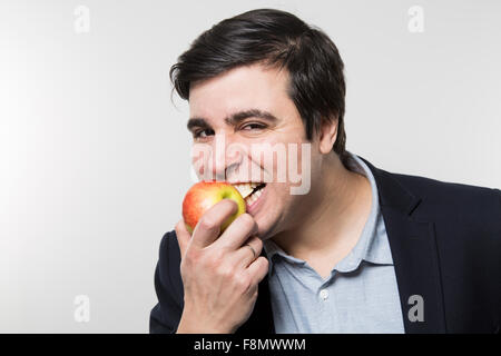 Dark-haired european businessman with a cheerful look takes a bite out of an small apple with his eyes opened while in front of  Stock Photo