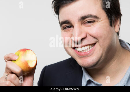 Dark-haired european businessman with a grin takes a bite out of an small apple with his eyes opened while in front of a gradien Stock Photo