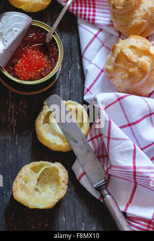 Home made Profiteroles cut and ready to stuff for red caviar with vintage knife and tin of red caviar on white kitchen towel ove Stock Photo