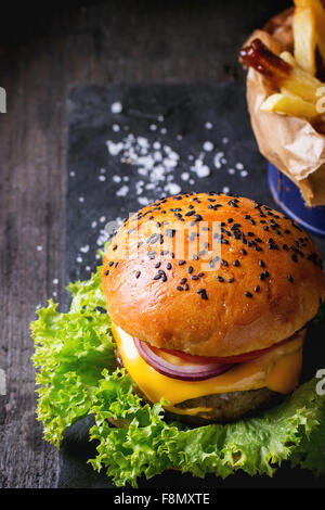 Fresh homemade burger with black sesame seeds on wooden cutting board with French fries, served with ketchup sauce in glass jar Stock Photo