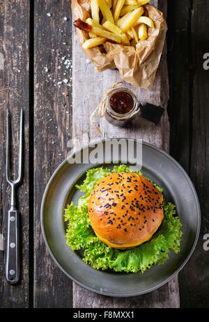 Fresh homemade burger with black sesame seeds and fried potatoes in backing paper, served with ketchup sauce in glass jar and me Stock Photo