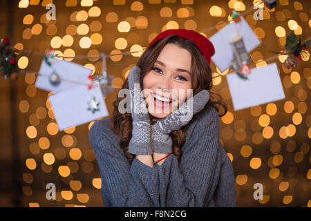 Lovely cheerful young woman in grey sweater and mittens posing over handmade decoration and glittering background Stock Photo