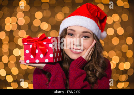 Dreaming smiling curly woman in santa claus hat holding gift on palm over glittering background Stock Photo