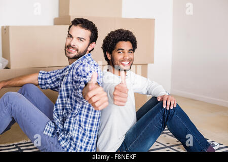 Happy gay couple sitting on floor back-to-back showing thumbs up Stock Photo