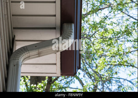 Aluminum gray downspout drain pipe leading down from brown eaves trough, with tree branches and leaves in blurred background. Stock Photo