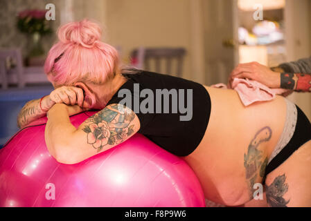 Woman in labour leaning on a yoga ball and having an ice pack held to her back by her partner at an unassisted home birth Stock Photo