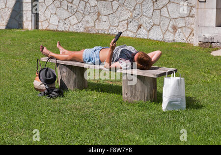 tanned middle age caucasian woman in shorts and sunglasses lying after shopping on a bench outdoor and reading electronic book Stock Photo