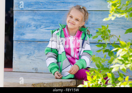 Six year old girl sitting on the porch of a wooden house and smiling happily Stock Photo