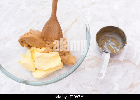 Wooden spoon mixing together smooth peanut butter and butter. Dirty measuring cup sits beside bowl on kitchen counter. Stock Photo