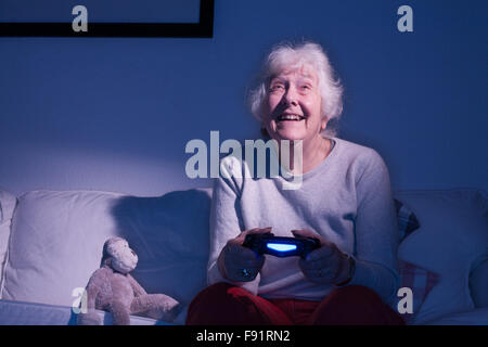 Elderly lady sitting on a sofa playing on a Playstation 4 next to a stuffed toy monkey. Stock Photo