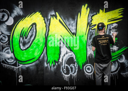 A rear view of a young teenage man spray painting street art - the word 'One' on a wooden hoarding wall UK