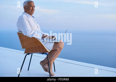 senior man working on laptop computer and looking ocean view sunset on balcony of modern home Stock Photo
