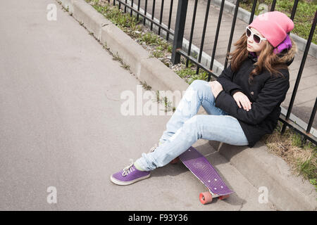 Teenage girl in jeans and sunglasses sits on her skateboard near urban fence Stock Photo