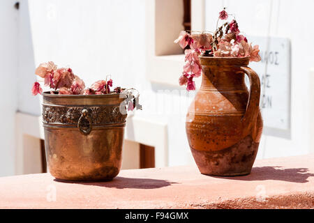 Cyclades, Santorini, Thira. Oia. Copper une, and orange coloured clay jug, side by side, both with wilted pink flowers in, on reddish wall. Stock Photo