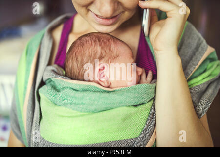 Newborn baby hold by mother in the baby wrap carrier. Mum calling at the phone. Stock Photo