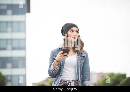 Half length of young handsome caucasian blonde straight hair woman walking outdoor in the city, holding a smartphone, overlooking left, pensive - social network, communication, technology concept Stock Photo