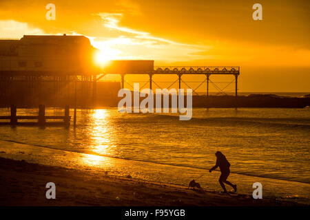 Aberystwyth, Wales, UK. 14th December, 2015.   In a short break between boughts of heavy rain, a woman walks her dog on the beach, as behind her the sun sets dramatically  over the silhouette of Aberystwyth's pier    Credit:  keith morris/Alamy Live News Stock Photo