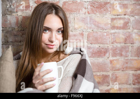 Happy woman in warm plaid holding cup with hot tea over brick wall background and looking at camera Stock Photo