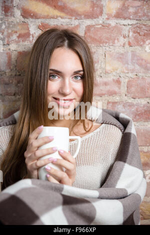 Smiling woman in warm plaid holding cup with hot coffee over brick wall background and looking at camera Stock Photo