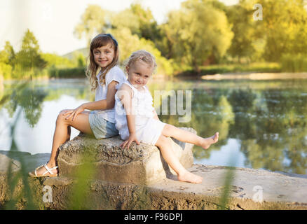Two cute little sisters sittting by the lake Stock Photo