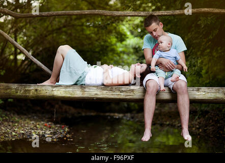 Happy family with little baby boy is sitting on the bridge over the river Stock Photo