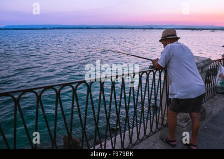 Old man fishing, rear view at dusk of a senior man fishing alone along the esplanade on Ortigia island, Syracuse (Siracusa), Sicily. Stock Photo