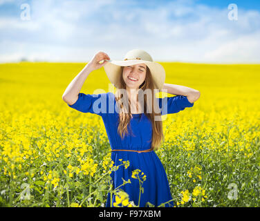 Happy young girl in blue dress and straw hat enjoying free time in yellow colza field Stock Photo
