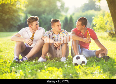Three happy friends spending free time together in park sitting on grass, drinking beer and chatting Stock Photo