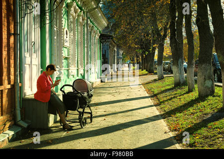 Woman with a baby in a pram sits on a wooden box and uses her mobile phone on an autumnal footpath in Ufa Russia during Stock Photo