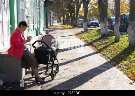 Woman with a baby in a pram sits on a wooden box and uses her mobile phone on an autumnal footpath in Ufa Russia during Stock Photo