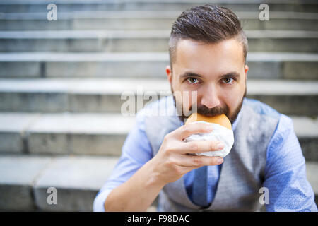 Modern hipster businessman having lunch break, sitting on stairs in town center and eating burger Stock Photo