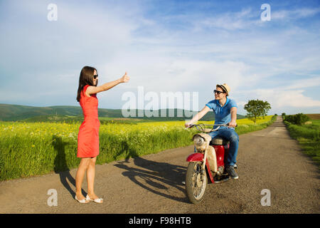 Beautiful young woman is hitch-hiking on the read near the fild. Young handsome man on retro motorbike is stopping to pick her u Stock Photo