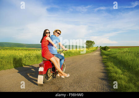 Happy young couple in love on retro motorbike driving togetger and ejoying the trip in green field Stock Photo