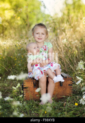 Summer outdoor portrait of cute little girl holding her baby sister and sitting on old suitcase Stock Photo