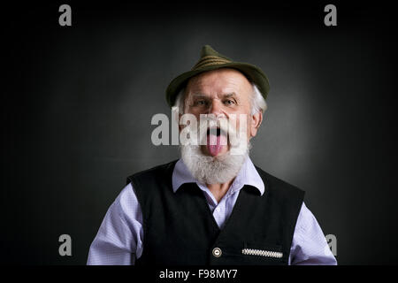 Portrait of old bearded bavarian man in traditional felt hat, sticking his tongue out in studio on black background Stock Photo