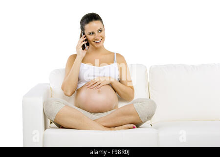 Studio portrait of pregnant woman with smartphone sitting on sofa isolated on white background Stock Photo