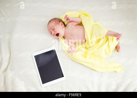 Crying newborn baby girl lying on bed next to digital tablet Stock Photo