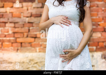 Outdoor portrait of beautiful pregnant woman in white dress holding her belly, brick wall in background, Detail of hands and bel Stock Photo