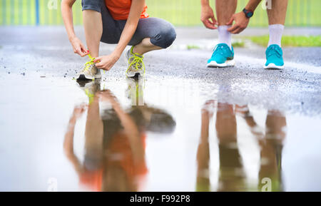 Young couple tying laces on asphalt sports field in rainy weather. Details of legs and sports shoes. Stock Photo