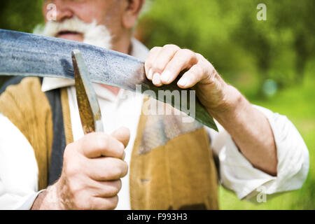 Old farmer with beard sharpening his scythe before using to mow the grass traditionally Stock Photo