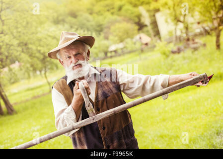 Old farmer with beard preparing his scythe before using to mow the grass traditionally Stock Photo