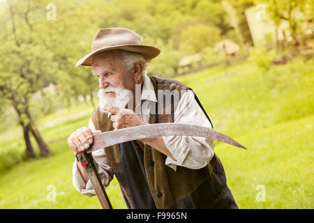 Old farmer with beard preparing his scythe before using to mow the grass traditionally Stock Photo