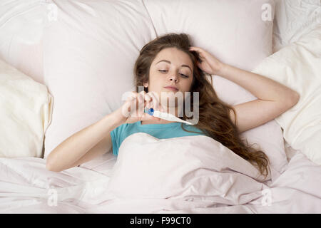 Sick woman with thermometer is lying in bed. She has cold, flu and high fever. Stock Photo