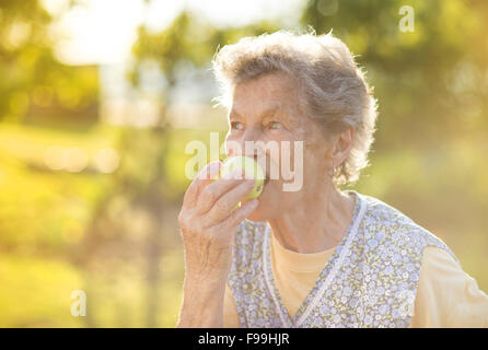 Portrait of senior woman in apron eating apple in the sunny garden Stock Photo