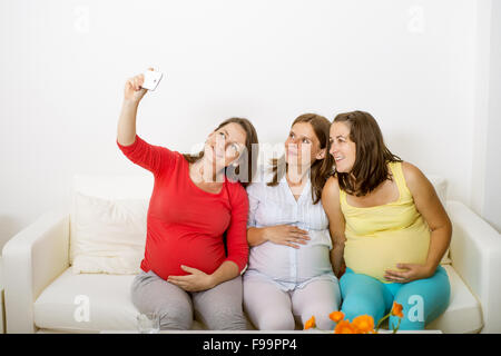 Three pregnant women sitting on sofa, smiling and taking selfie Stock Photo