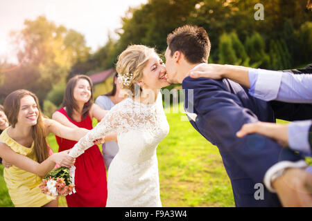 Funny portrait of newlywed couple kissing, bridesmaids and groomsmen pulling them away in green sunny park Stock Photo