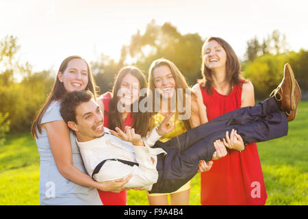 Funny portrait of bridesmaids holding groom on their hands in green sunny park Stock Photo