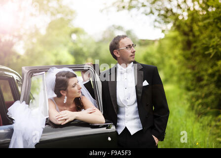 Beautiful happy young bride and groom posing by the retro car. Outdoor wedding portrait. Stock Photo